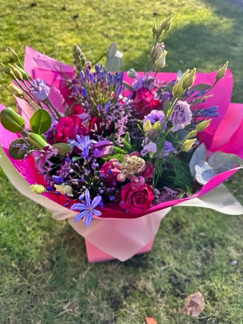 A floral arrangement with blue and pink flowers, including blue agapanthus and pink roses, tied together and presented in a pink wrapping with a aqua of water visible at the base.