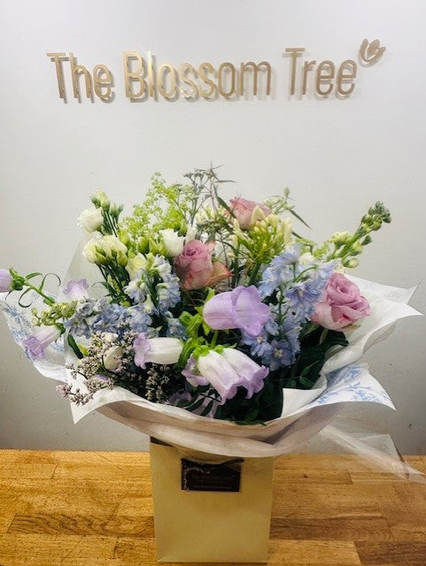 A hand tied aqua bouquet with a variety of flowers in blue, purple, and white tones, arranged loosely with greenery, presented in eco-friendly wrapping with a gift tag.