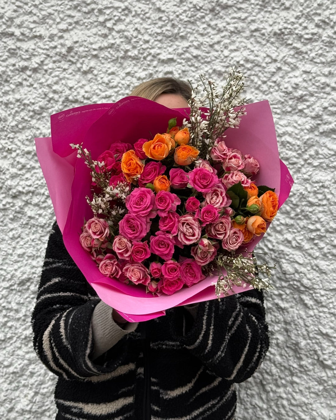 Person holding a bouquet of pink and orange flowers wrapped in pink paper against a textured white wall.