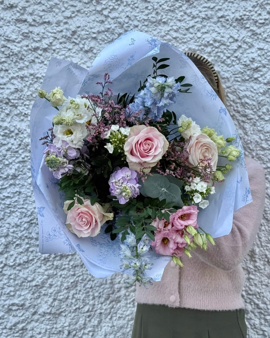 Bouquet of Pink, Purple, White and Blue flowers held by a person against a textured wall.