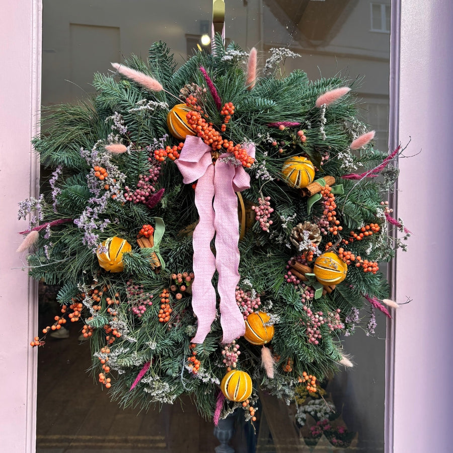 Pink Christmas Wreath, vibrant orange ilex berries, delicate pink pepperberries, and soft limonium, complemented by whole oranges and wispy dried bunny's tail for texture.
