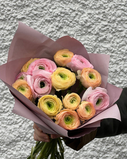 Bouquet of pink, yellow, and orange Ranunculus flowers wrapped in pink paper against a textured grey background.