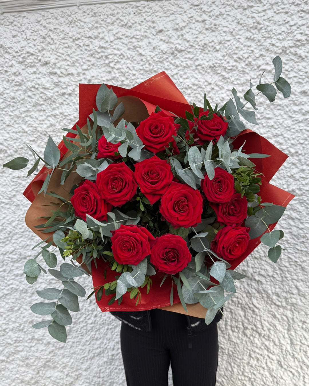 Bouquet of red roses with greenery wrapped in red paper against a white wall.