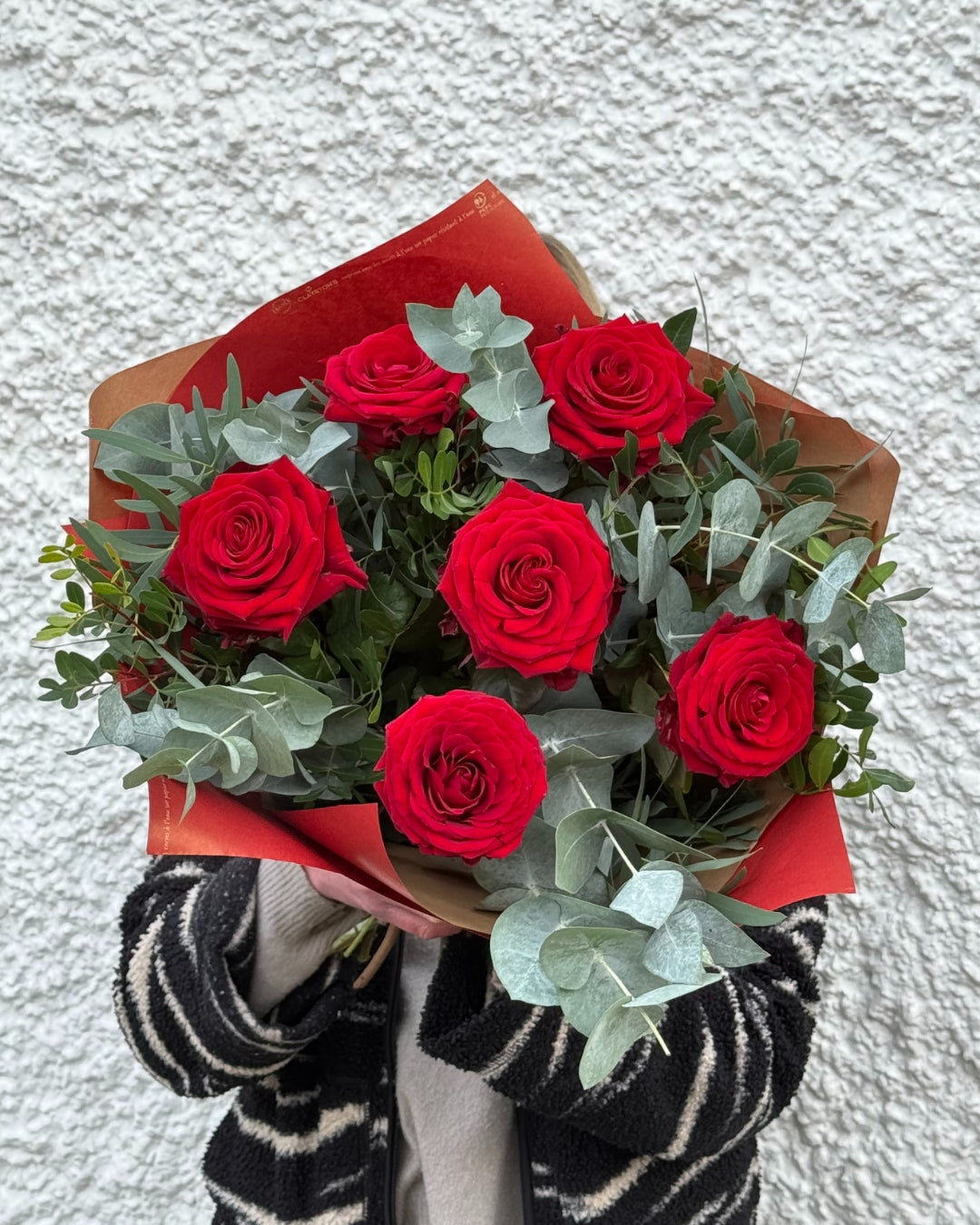 Bouquet of red roses with greenery held by a person against a light grEy background.