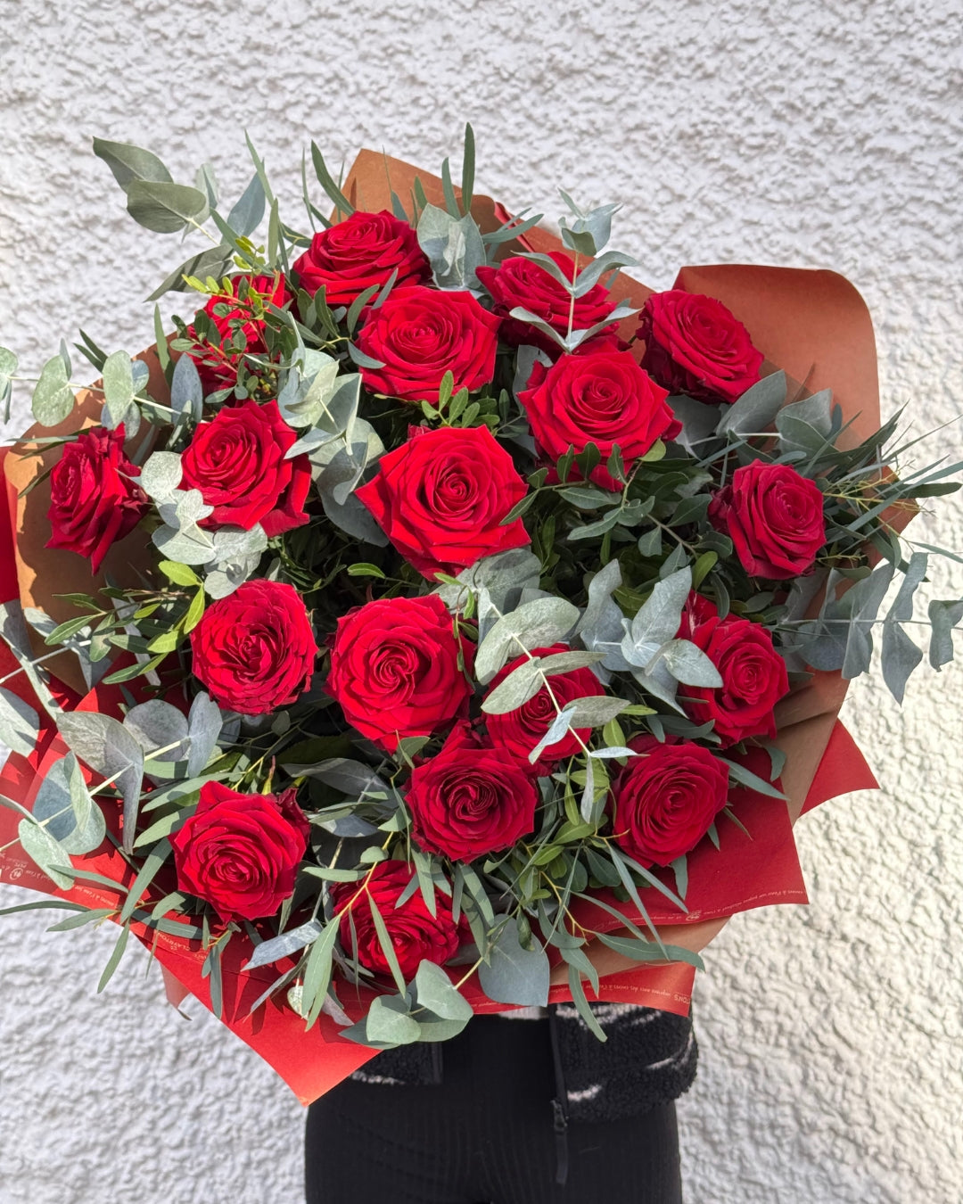 Bouquet of 18 red Naomi Roses with greenery against a textured white wall