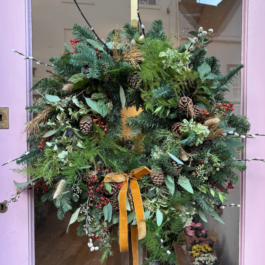 Decorative wreath with greenery, berries, fir cones, blue scottish thistle, dried bunnies tail, pussy willow and a yellow ribbon on a pink door.