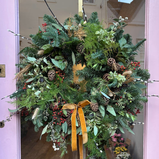 Decorative wreath with greenery, berries, fir cones, blue scottish thistle, dried bunnies tail, pussy willow and a yellow ribbon on a pink door.