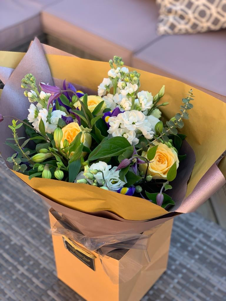 A bouquet of flowers including yellow roses, white and purple flowers, and greenery, wrapped in brown paper.