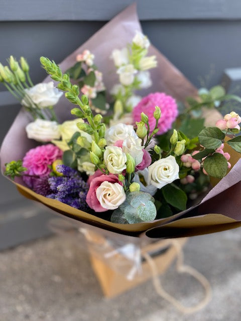 A wrapped bouquet of assorted fresh flowers including pink roses and purple Statice and Dahlias arranged in a seasonal selection.