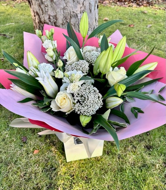 A bouquet of white and green flowers including roses, lilies, and other foliage, wrapped in pink paper and presented in front of a tree.